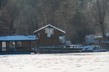 Winter landscape in Kyiv, with a floating houseboat or cabin on a lake, snow-covered roof and surroundings, muted colors, soft glow, long shadows, horizontal distort Kyiv, Ukraine - February 16, 2025