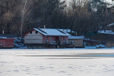 Winter scene in Kyiv, featuring a rustic cabin on frozen lake with boat dock Soft color palette of white, grey, brown, and red objects Overcast sky suggesting dawn Kyiv, Ukraine - February 16, 2025