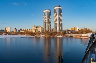 Kyiv şehrinin kış manzarası, donmuş bir su kütlesinden bakıldığında, silindirik çekirdekli iki modern gökdelen, cam yüzeylerine yansıyor, Kyiv, Ukrayna - 16 Şubat 2025
