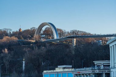 Dinyeper Nehri ile Kiev manzarası, günbatımında şehir manzarası, tepede modern mimari, beyaz kemerli eğri köprü benzeri yapı, gri sütunlar, Kyiv, Ukrayna - 19 Ocak 2025