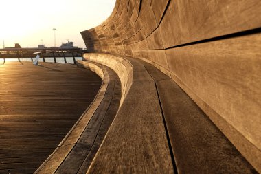 Gün batımında tahta kaldırımdaki eğimli bir bankın yakın görüntüsü, ahşabın dokusunu ve ılık altın ışığı gözler önüne seriyor.