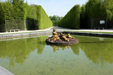 Bereketli yeşillik ve yansıtıcı gölle çevrili heykellerle dolu bir çeşme içeren sakin bir bahçe sahnesi.