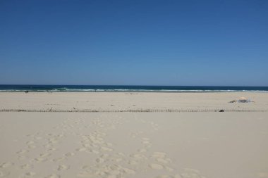 Berrak mavi gökyüzü, yumuşak dalgalar ve kumdaki ayak izleriyle sakin bir sahil sahnesi.