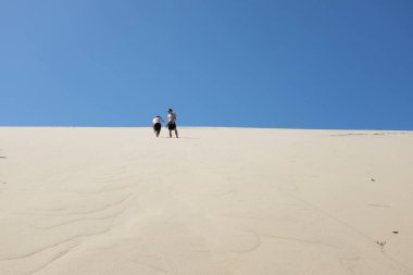 İki insan açık mavi gökyüzünün altında kumlu bir tepeye tırmanıyor.