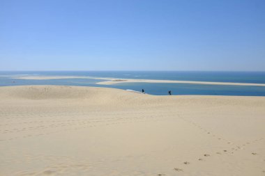 Altın kum tepecikleri, sakin mavi bir deniz ve kıyıda yürüyen iki insanın olduğu berrak bir gökyüzü içeren sakin bir sahil manzarası.