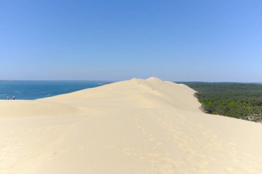 Mavi bir okyanusa bakan engin kumlu bir manzara, berrak bir gökyüzü ve çok az insan uzaklarda yürüyor.