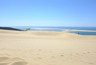 Altın kum tepelerinin, berrak mavi gökyüzünün ve arka planda sakin bir okyanusun olduğu sakin bir sahil manzarası.