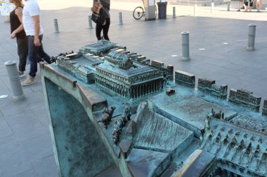 Tarihi bir binanın detaylı bronz modeli, mimari özellikler sergileniyor, şehrin arka planında yürüyen insanlar var.