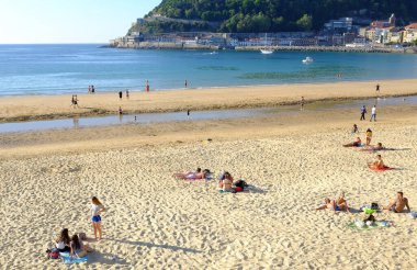 Güneşli bir sahil sahnesi, kumsalda dinlenen insanlar, biraz güneşlenen, diğerleri kıyıda yürüyen, ve arka planda sakin bir deniz.