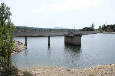 Berrak bir gökyüzünün altında yeşil ve tepelerle çevrili sakin bir göle uzanan beton bir yapıya sahip huzurlu bir baraj manzarası.