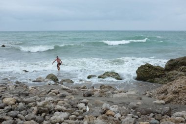 Okyanusta yürüyen bir adam kayalık kıyılarda, dalgalar çarpıyor, bulutlu gökyüzü