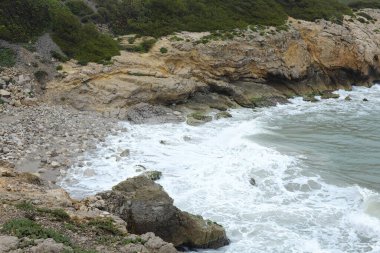 Sahile vuran dalgalarla dolu kayalık bir kıyı şeridi, arka planda yemyeşil bir zemin ve kumsalda çakıl taşları ve kayaların karışımı.