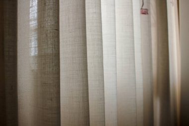 Parlak bir oturma odasında asılı şık pencere perdelerinin iç çekimi. Görüntü, kumaşın dokusunu ve pencereden süzülen yumuşak doğal ışığı gösteriyor. Sıcak ve havadar bir iç atmosfer yaratıyor..