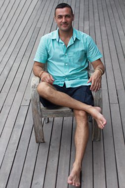 Tabii ve sağlam. Güzel genç adam ahşap bir sandalyede oturan ve bir arka plan üzerinde ahşap zemin ile bir odaya bakıyor