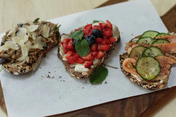 Bruschettas mit rotem Fisch und einer Gurke, Banane mit Erdnussmark und Mandeln, Tomate mit einer Gurke, Bruschettas auf einem Holzbrett, leckeres Essen — Stockfoto