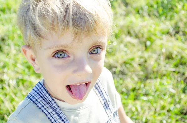 Lindo niño rubio mostrando su lengua, jugando en el campo de hierba verde en el soleado día de primavera . — Foto de Stock