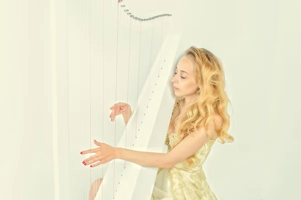 Elegante mujer en vestido dorado con pelo largo y rubio tocando el arpa, sobre fondo blanco — Foto de Stock