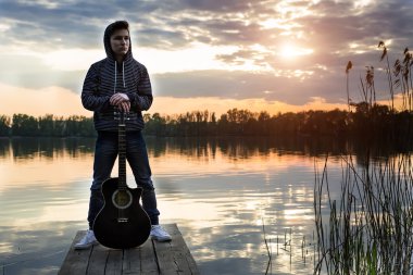 Köprüde onun gitar ile hood ayakta karşı bir gün batımı zemin Nehri üzerinde akşamları genç adam