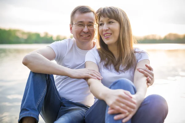 Pareja sonriente sentada en el puente a orillas del lago al atardecer —  Fotos de Stock