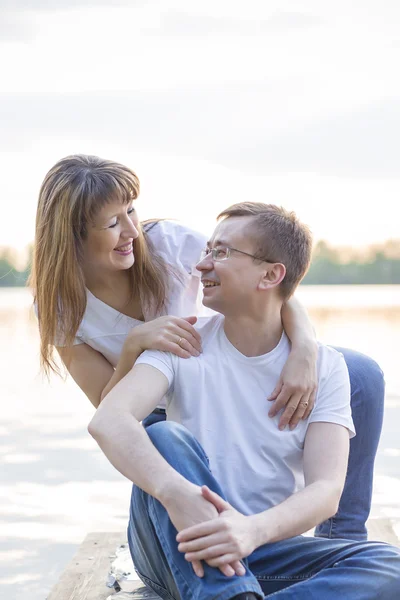 Pareja sonriente sentada en el puente a orillas del lago al atardecer —  Fotos de Stock