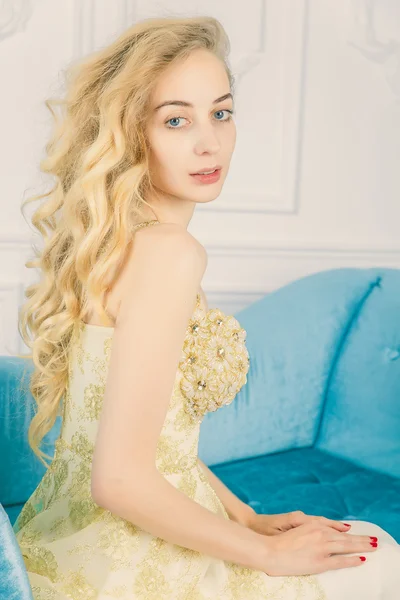 Portret van een mooie jonge blonde in een lang gouden jurk op de Bank. Studio. Haute Couture. Emoties — Stockfoto