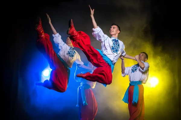 Чотири молодих людей в національних українських костюмах танцюють на темному тлі з димом — стокове фото