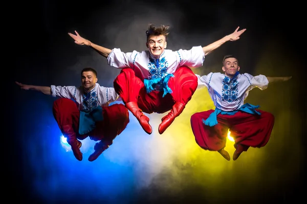Трьох молодих людей в національних українських костюмах танцюють і стрибати на темному тлі з димом — стокове фото