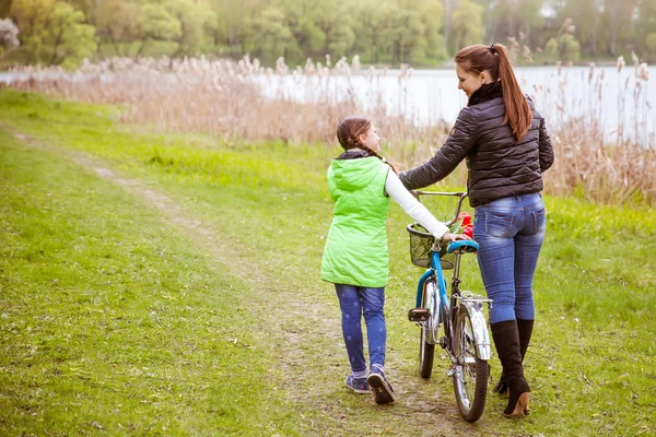 Hija y madre caminan a lo largo de la orilla del lago con una bicicleta y hablan. Valores familiares, educación — Foto de Stock