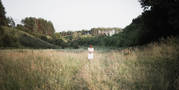 Young brunette girl with bouquet in white dress stands in the middle of the field with his head down. Sadness, loneliness, doubt. Retro photo processing