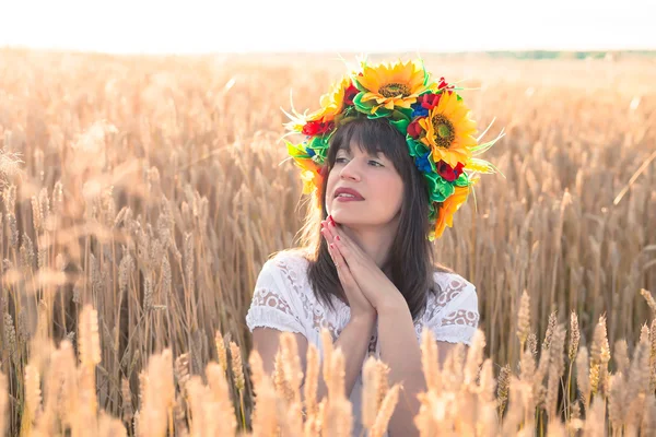 Jonge vrouw in een krans van zonnebloemen gevouwen zijn handen in de buurt van het gezicht en zit in de tarwe — Stockfoto