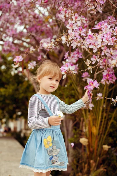 Маленька дівчинка в смугастій джинсовій сукні, що стоїть біля квітучого — стокове фото