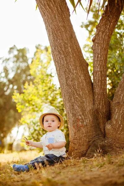 Büyük gözlü küçük küçük çocuk şapkalı bir ağacın yanında günbatımında oturur. — Stok fotoğraf