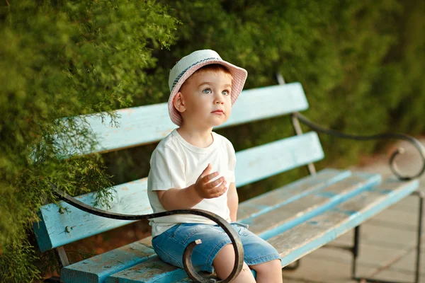 Pequeño niño en un sombrero sentado en un banco en el verano y lo — Foto de Stock