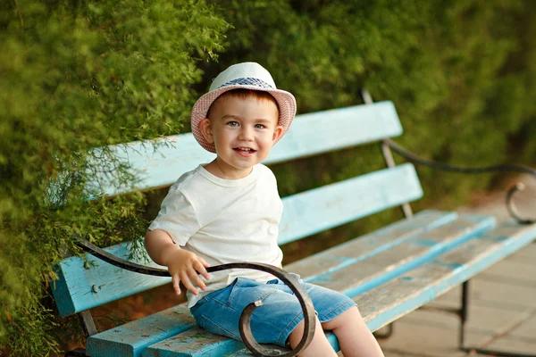 Pequeño niño en un sombrero sentado en un banco y sonriendo en el s —  Fotos de Stock