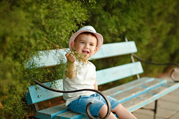 Pequeño niño en un sombrero sentado en un banco y sonriendo en el s —  Fotos de Stock