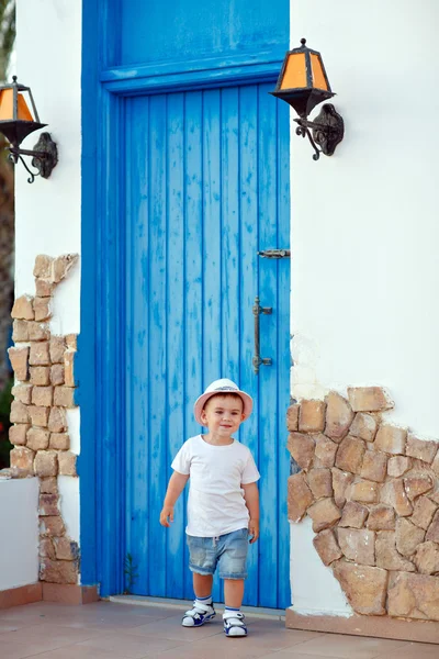 青いドアの家のそばに立っている帽子をかぶった小さな男の子 — ストック写真