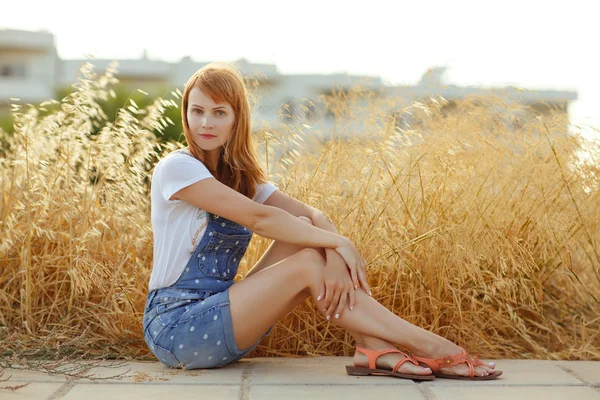 Affascinante ragazza dai capelli rossi in tuta di jeans seduta su un backgrou — Foto Stock