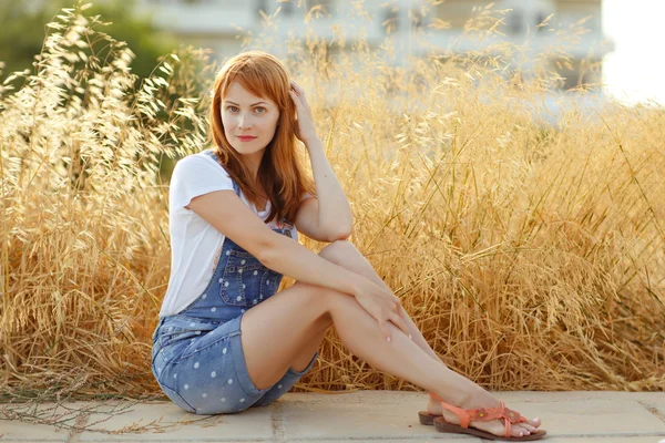 Affascinante ragazza dai capelli rossi in tuta di jeans seduta su un backgrou — Foto Stock