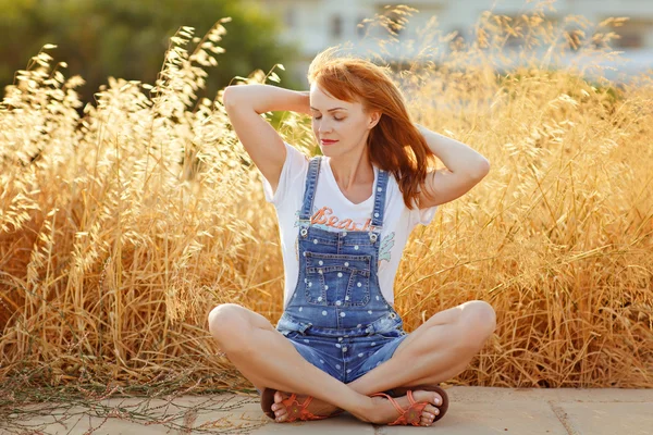 Menina ruiva encantadora em macacão jeans sentado em um backgrou — Fotografia de Stock