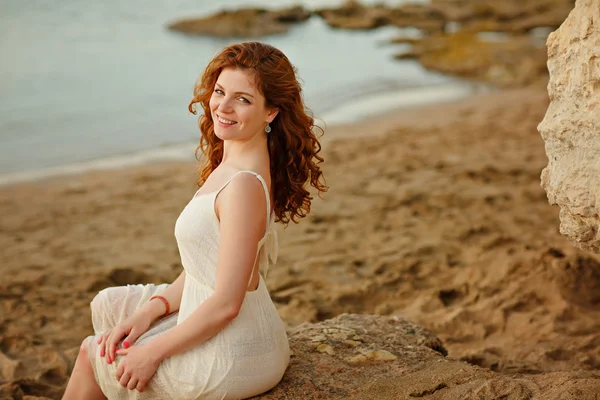 La ragazza sensuale dai capelli rossi in un vestito bianco sorride e si siede su — Foto Stock