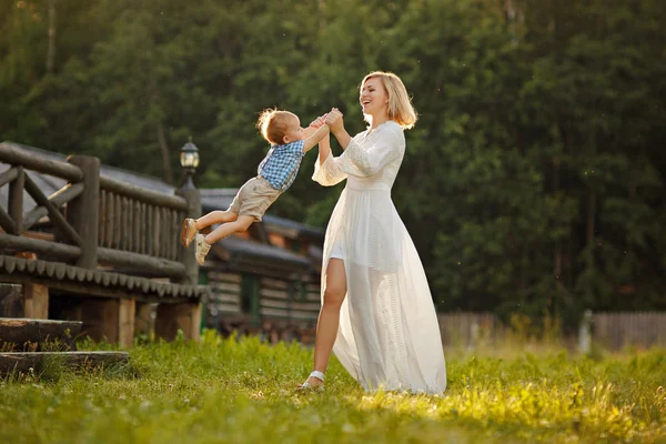 Mamá rubia en vestido blanco ríe y gira hijo al atardecer en summ — Foto de Stock