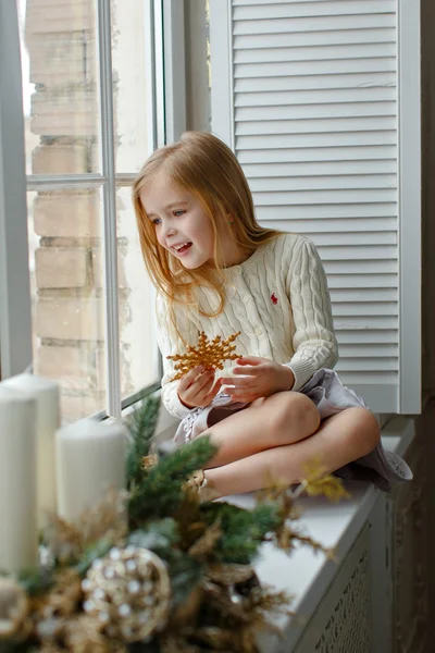 Vacker liten blond flicka med blå ögon sitter på fönstret — Stockfoto