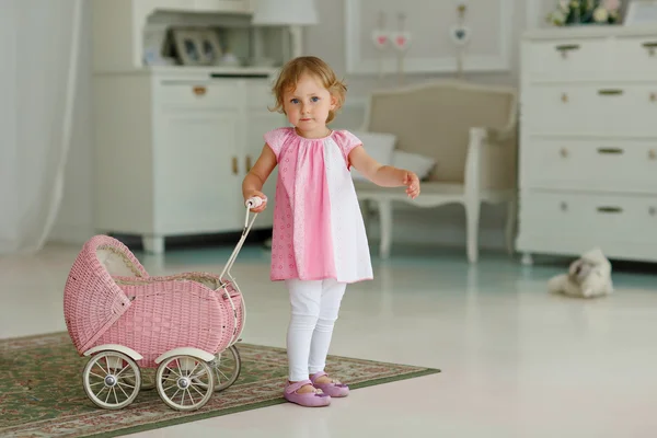 Pequena menina bonita em um vestido rosa brinca com um carrinho de bebê — Fotografia de Stock