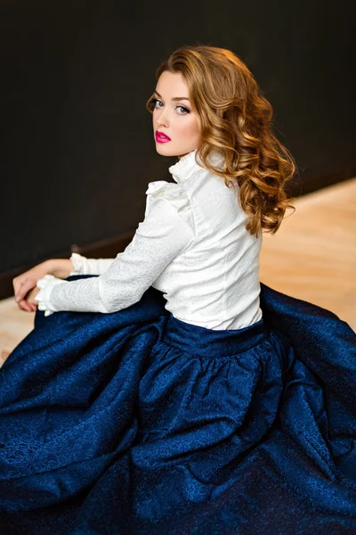 Retrato de una chica sensual glamorosa de pelo rojo muy hermosa con una blusa blanca y falda azul, sentada en el suelo en el estudio sobre un fondo oscuro —  Fotos de Stock