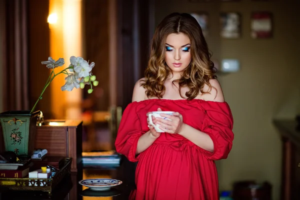 Портрет красивой чувственной девушки с закрытыми глазами в красном платье, держащей чашку в интерьере — стоковое фото