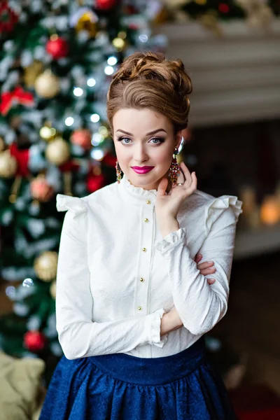 Chica rubia muy hermosa en una falda azul y blusa blanca de pie alrededor de un árbol de Navidad y la chimenea y sonrisas — Foto de Stock