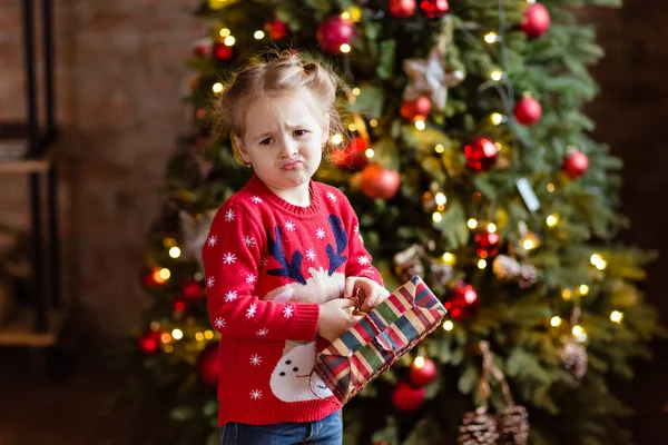 Niña niña en el suéter rojo sosteniendo un regalo y hace la cara —  Fotos de Stock