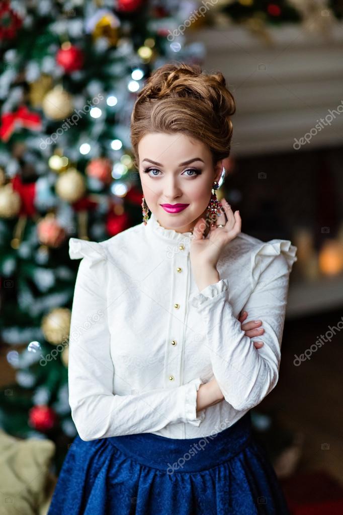 Very beautiful blonde girl in a blue skirt and white blouse standing around a Christmas tree and the fireplace and smiles