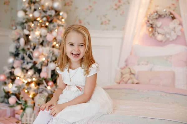 Muy linda niña rubia en un vestido blanco sosteniendo una caja de regalo sobre un fondo de árboles de Navidad en el interior de la casa — Foto de Stock