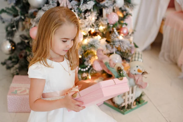 Muy linda niña rubia en un vestido blanco sosteniendo una caja de regalo sobre un fondo de árboles de Navidad en el interior de la casa — Foto de Stock